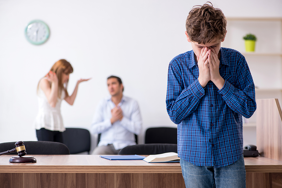 Mother and father arguing in the background while a teenage boy in a blue shirt holds his face over his hands. 