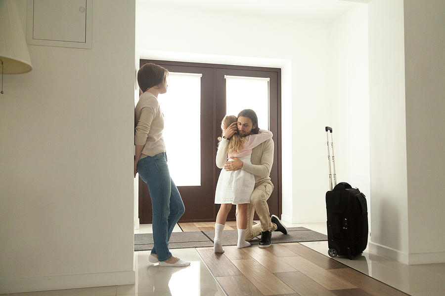 Little girl hugging her dad in front of the door, next to a packed suitcase. 