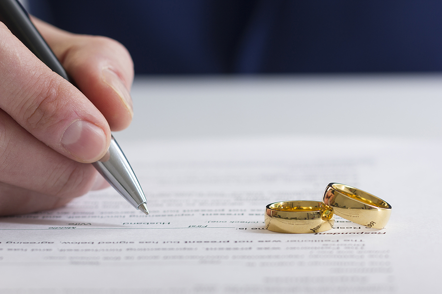 Close up of a hand signing divorce decree with two gold wedding bands on the document. 